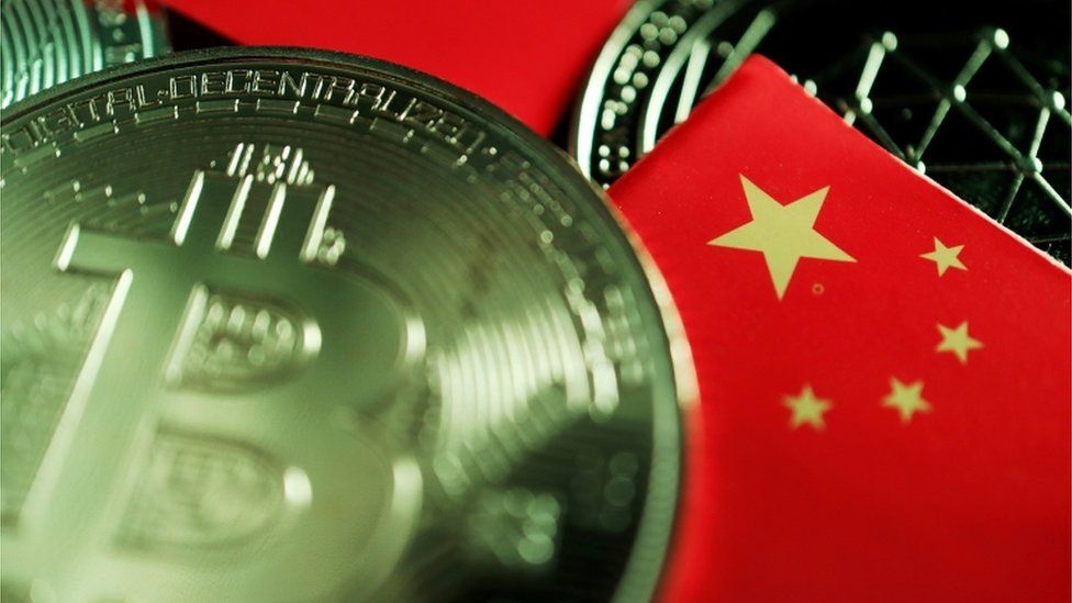 A real-world coin designed with the Bitcoin logo lies scattered on a table amid some miniature Chinese flags